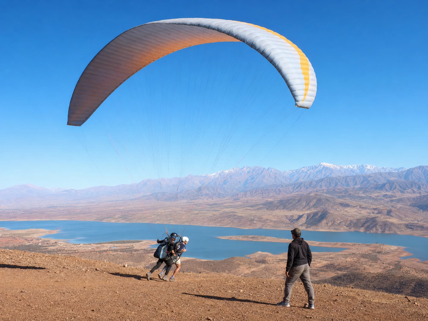 A helmeted tandem paraglider launching above Lake Lalla Takerkoust with the High Atlas beyond